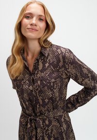 Koszula-dress w brązowym i beżowym wzorze w węża, z długimi rękawami, dwiema kieszeniami na piersi oraz paskiem w talii, który pozwala na regulację dopasowania.