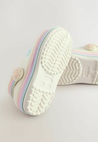 Scarpe bianche con solette striate arcobaleno mostrate a testa in giù, che rivelano suole testurizzate e decorazioni a forma di fiore con la scritta "Fatto con amore".