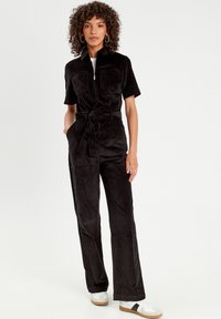 Black corduroy jumpsuit with short sleeves, two chest pockets, a front zipper, and a belt at the waist. Styled with white sneakers.