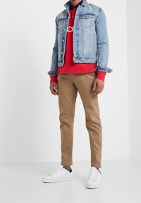 Veste en denim bleu clair avec poches à boutons, sweatshirt rouge en dessous, pantalons kaki et baskets blanches avec accents noirs.