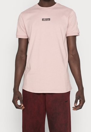 Mężczyzna w jasnoróżowej koszulce z krótkim rękawem z logo "ellesse" na piersi oraz w bordowych szortach na białym tle.