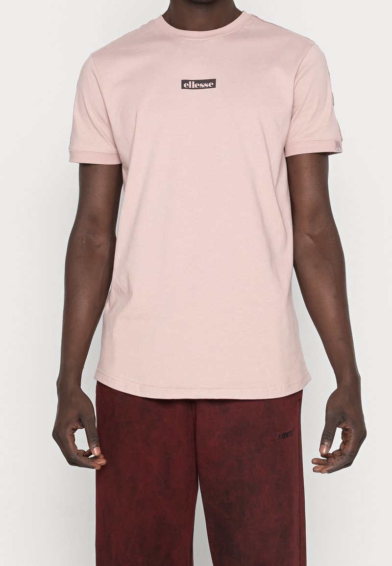 Homme portant un t-shirt à manches courtes rose clair avec le logo "ellesse" sur la poitrine et un short rouge foncé, sur fond blanc.
