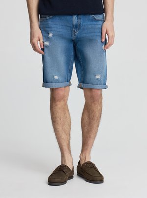Gambe maschili che indossano pantaloncini di denim blu con risvolti e toppe consumate, abbinati a mocassini marroni, in piedi davanti a uno sfondo semplice.