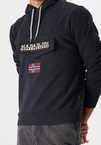 Osoba ubrana w granatowy hoodie marki Napapijri z przednią kieszenią i naszywką z flagą Norwegii, w zestawie z szarymi dżinsami.