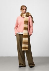 Różowy, puszysty sweter o luźnym kroju; długa, paskowana chusta w kolorze beżowym i brązowym; szerokie, oliwkowe spodnie; czarne buty.