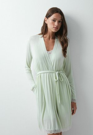 Jeune femme aux longs cheveux bruns portant une robe de chambre vert clair nouée à la taille avec une bordure en dentelle aux manches et à l'ourlet, debout contre un mur blanc.