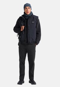 Schwarze isolierte Weste über einem schwarzen Langarmhemd, kombiniert mit schwarzen Hosen und dunklen Schuhen. Eine blaue Mütze und ein Schal runden den Look ab.