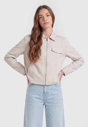 Jonge vrouw met lang golvend haar draagt een lichtbeige jas met rits en lichtblauwe wijde spijkerbroek, staand met handen in de zij.