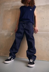 Navy sleeveless knit vest with a v-neck and logo patch, paired with loose blue denim jeans and black sneakers with white laces.
