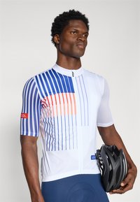 Short-sleeve cycling jersey in white with blue and orange vertical stripes, textured fabric, front zipper, and logo accent. Black helmet held in hand.