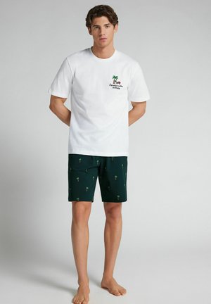 Jonge man staat blootsvoets, draagt een wit T-shirt met een afbeelding van een palmboom en een auto, en een donkere korte broek met kleine patronen van palmboom.