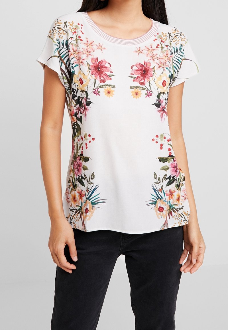 Blouse blanche à manches courtes avec des motifs floraux colorés sur les épaules et sur les côtés, portée avec un pantalon noir, se tenant devant un fond uni.