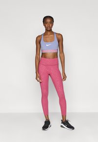 Αθλητικό σουτιέν Nike Pro σε ανοιχτό μωβ χρώμα με λευκό λογότυπο, συνδυασμένο με ροζ κολάν ψηλόμεσο με υφή, ολοκληρωμένο με μαύρα αθλητικά παπούτσια.