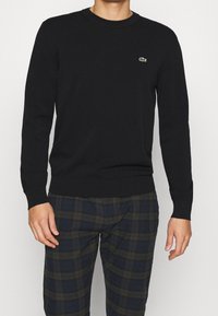 Homme portant un pull noir à manches longues avec un petit logo de crocodile vert et un pantalon à carreaux bleu foncé et vert.
