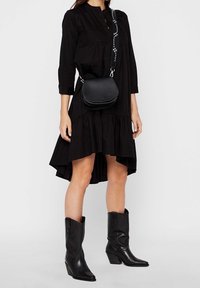 Vestido negro de manga larga con dobladillo acampanado, combinado con un bolso cruzado redondo negro y botas de vaquero negras hasta el tobillo. Textura de tela suave.