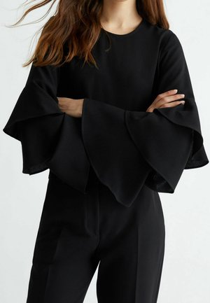 Femme aux longs cheveux bruns portant un haut noir avec de grandes manches évasées superposées et un pantalon noir assorti, les bras croisés sur la poitrine.