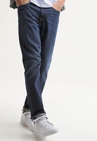 Mörkblå denimjeans med rak skärning och ton-i-ton-sömmar, tillsammans med vita sneakers. Halen är lätt uppvikt, vilket visar fållen.