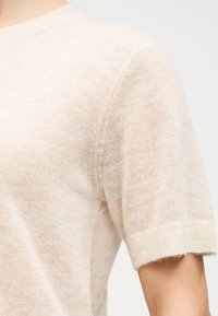 Μαλακό μπεζ πλεκτό πουλόβερ με κοντά μανίκια, φορεμένο στο πάνω μέρος του σώματος, παρουσιάζει λεπτομέρειες στους ώμους και τα μπράτσα με λεπτή υφή υφάσματος.