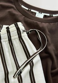 Zbliżenie na brązową bluzę z okrągłym dekoltem oraz białe spodnie z pionowymi brązowymi paskami i elastycznym pasem ze sznurkiem.