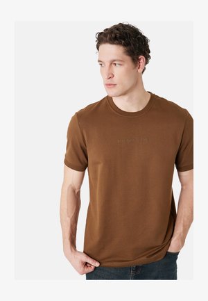 Man draagt een bruin shirt met korte mouwen met de tekst "ECHO THROUGH TIME", kijkt naar links, hand houdt de zoom van het shirt vast, tegen een effen achtergrond.