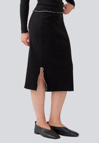 Jupe midi noire à côtes avec un ourlet à volants et un détail d fente. Associée à des chaussures plates noires, mettant en valeur une silhouette élégante.