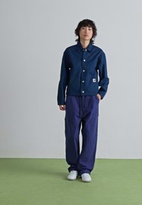 Personne debout sur un sol vert contre un mur gris, vêtue d'une veste navy avec pochette avant, de pantalons navy amples et de baskets blanches.