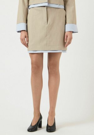 Kvinne iført beige skjørtedress med lyseblå mansjetter og svarte høye hæler, stående foran en ensfarget bakgrunn.