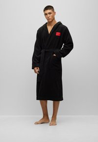 Black hooded bathrobe in soft fabric, featuring a front pocket and a red logo patch. The robe has a tie belt and knee-length design.