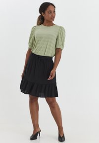 Top verde chiaro con trama e maniche a sbuffo, abbinato a una gonna nera con orlo arricciato. Modella che indossa tacchi neri.