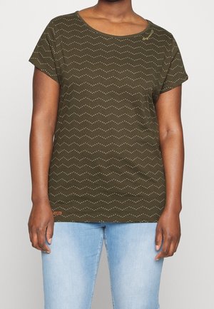 Woman wearing olive green short-sleeve top with white dotted wavy pattern and light blue jeans, standing against plain background.