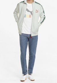 Lysegrøn zip-jakke med mørkegrønne skulderstriber, parret med en hvid T-shirt med et farverigt logo, blå jeans og hvide sneakers.