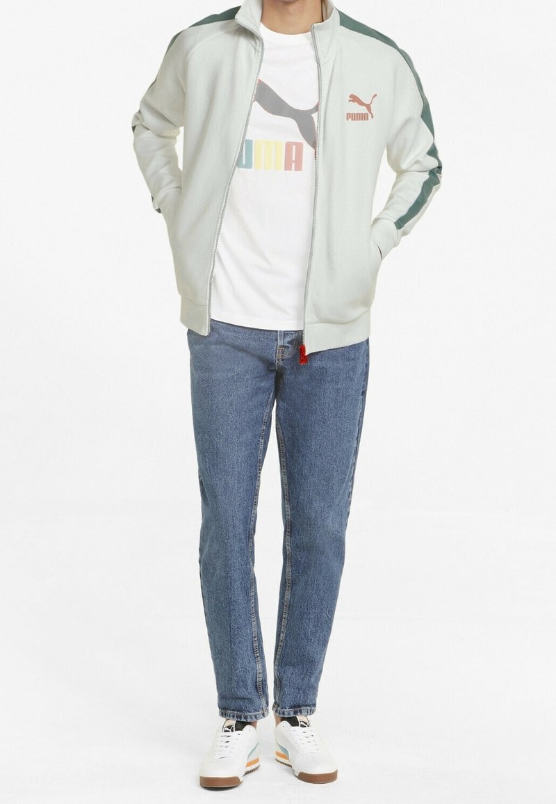 Lysegrøn zip-jakke med mørkegrønne skulderstriber, parret med en hvid T-shirt med et farverigt logo, blå jeans og hvide sneakers.