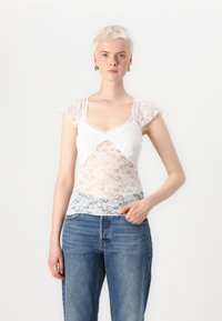 Hvitt blondertop med korte, fladdrende ermer, V-hals, og gjennomsiktige paneler over en solid grå blonderband i kanten, sammen med blå jeans.