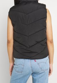 Schwarze ärmellose Steppweste mit Zickzacknähten, aus weichem Stoff gefertigt, mit hohem Kragen und glatter Textur.