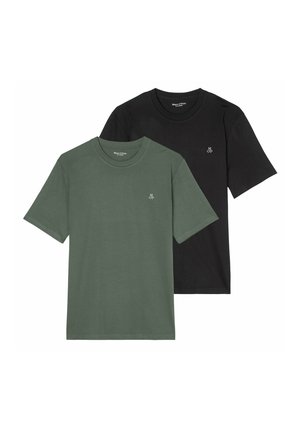 Joven con una camiseta negra lisa con un pequeño logo blanco "M O P" en el pecho y pantalones gris claro, con la mano en el bolsillo.