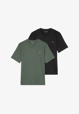 Giovane che indossa una maglietta nera semplice con un piccolo logo bianco "M O P" sul petto e pantaloni grigio chiaro, mano in tasca.
