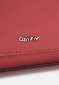Punainen tekstuurinen synteettinen materiaali, jossa on hopeinen "Calvin Klein" -logo selkeästi esillä. Reunojen ommellut yksityiskohdat lisäävät kestävyyttä.