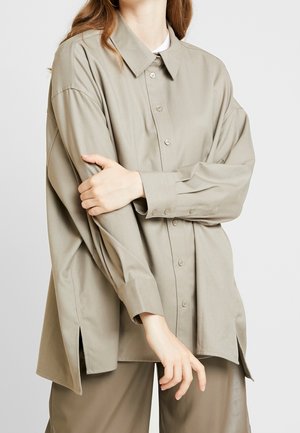 Personne portant une chemise beige ample boutonnée avec manches longues et fentes latérales, associée à un pantalon beige assorti, bras croisés.