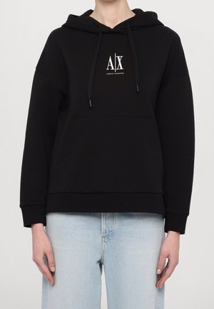 Personne portant un sweat à capuche noir Armani Exchange avec logo sur la poitrine et un jean bleu clair, debout devant un fond clair uni.