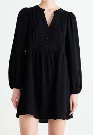 Czarna sukienka z długim rękawem, ze ściągaczem w talii, plisowanym przodem, dekoltem w kształcie litery V zapinanym na trzy guziki oraz lekko bufkami na ramionach, noszona przez osobę.