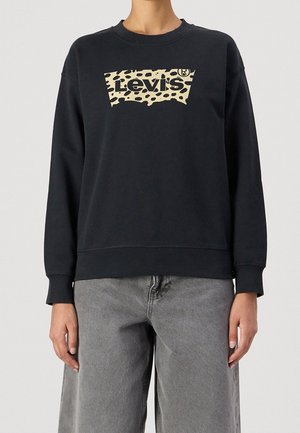 Personne portant un sweat-shirt noir Levi's avec un logo imprimé léopard beige, associé à un jean gris ample, visible du cou aux hanches.