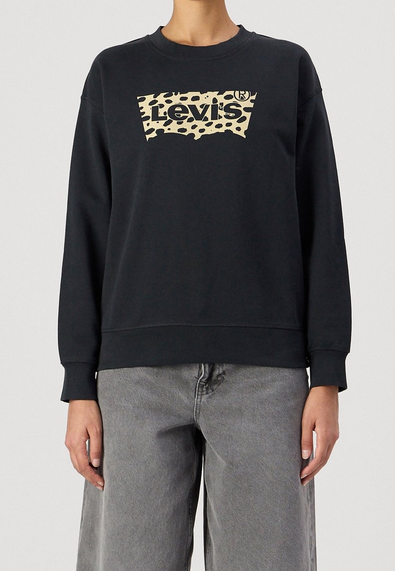 Personne portant un sweat-shirt noir Levi's avec un logo imprimé léopard beige, associé à un jean gris ample, visible du cou aux hanches.