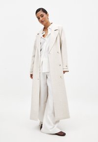 Abrigo largo beige con tejido texturizado, diseño de doble botonadura, grandes solapas y bolsillos laterales. Llevado sobre una camisa blanca y pantalones anchos.