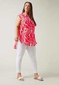 Rode mouwloze shirt met wit golvend patroon, knoopsluiting aan de voorkant en een hoge-lage zoom, gecombineerd met witte skinny jeans en witte sandalen.