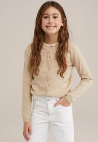 Bežs adīts kardigans ar sešiem pogām, apaļu dekoltē un garām piedurknēm, pārī ar baltām džinsām, kurām ir priekšējais kabata un jostas cilpa.