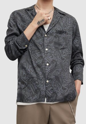 Osoba w ciemnej koszuli z wzorem paisley zapinanej na guziki, beżowe spodnie, srebrny naszyjnik, pierścionek i tatuaż na dłoni dotykającej kołnierza.