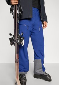 Ski hlače u živoj plavoj boji, s džepovima na patentni zatvarač i sivim detaljima na manžetama. Izrađene od izdržljivog, teksturiranog materijala za zimske sportove.