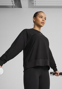 Black long-sleeve athletic sweatshirt with a rounded hem and ribbed cuffs, paired with black leggings, held with a silver dumbbell in one hand.