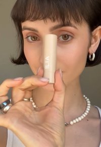 Applicateur ILIA en beige clair, texture lisse, forme cylindrique. Le modèle le tient près de lui tout en portant un collier de perles et des bagues.