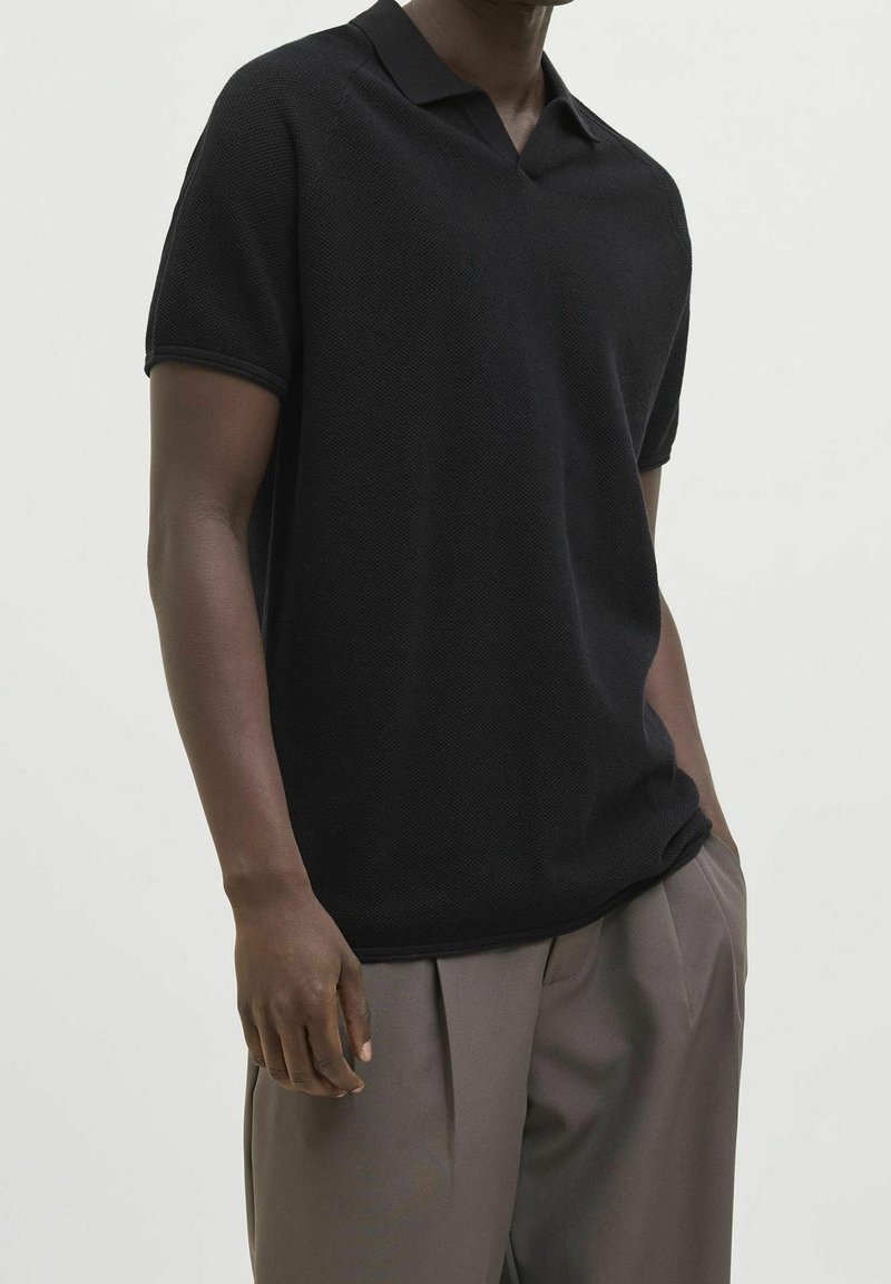 Polo noir à manches courtes en tissu texturé, doté d'un col et d'un décolleté en V. Associé à un pantalon gris clair.